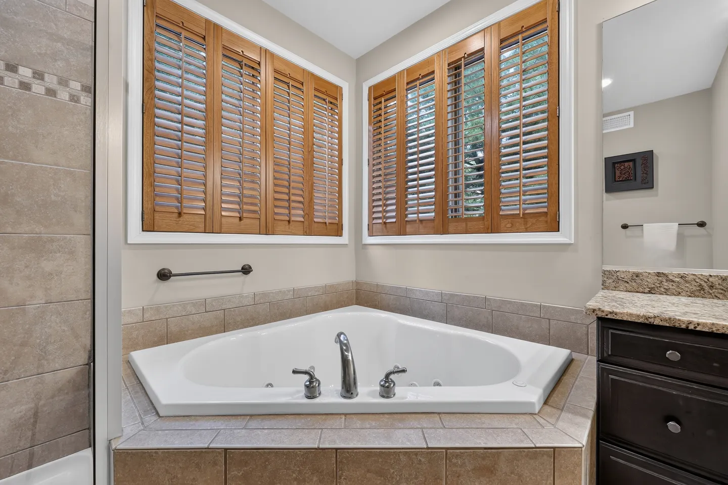 A corner soaking tub set into tile beneath two windows with wooden plantation shutters.