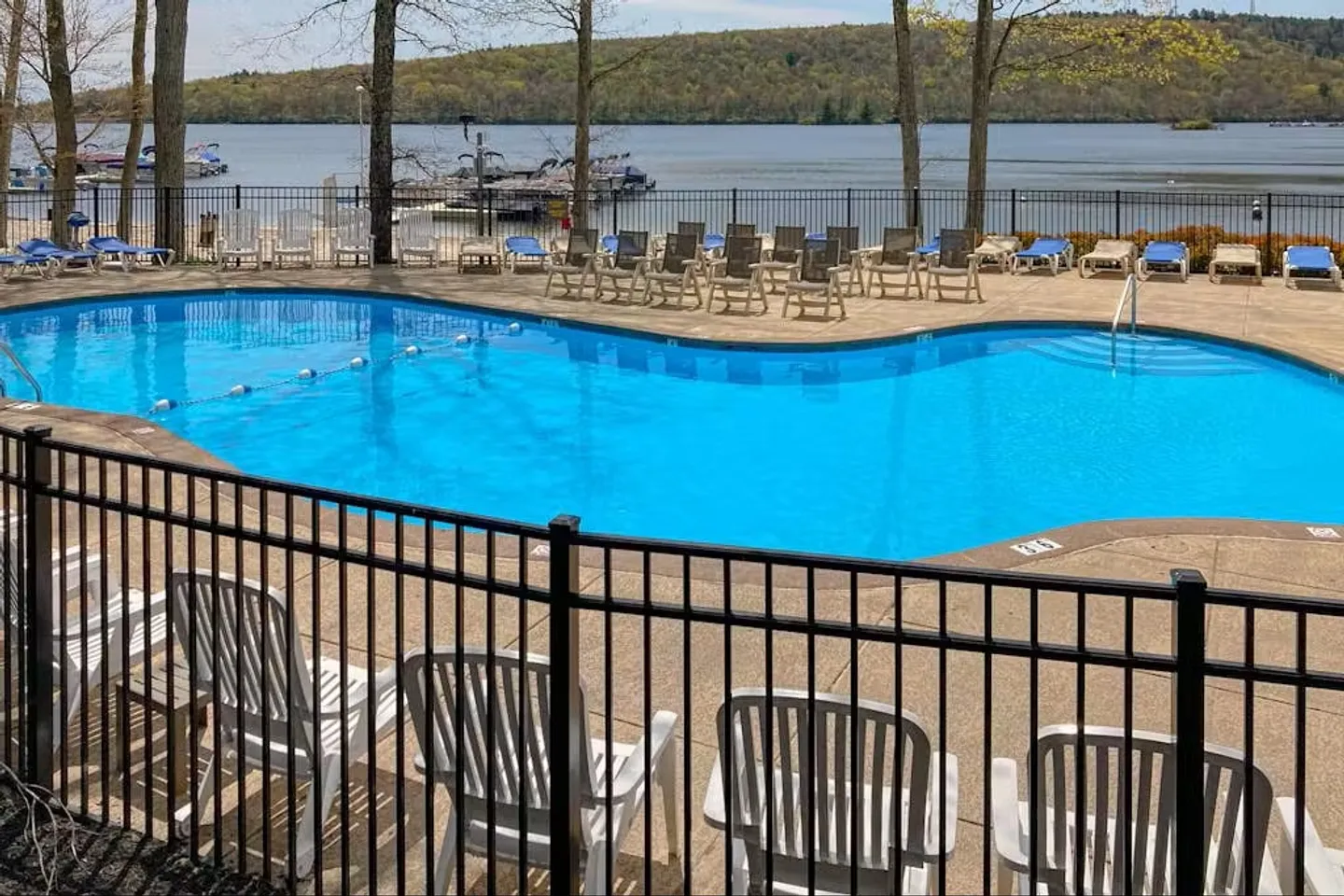 Fenced outdoor swimming pool with surrounding lounge chairs overlooking a lake with boat docks and forested hills.