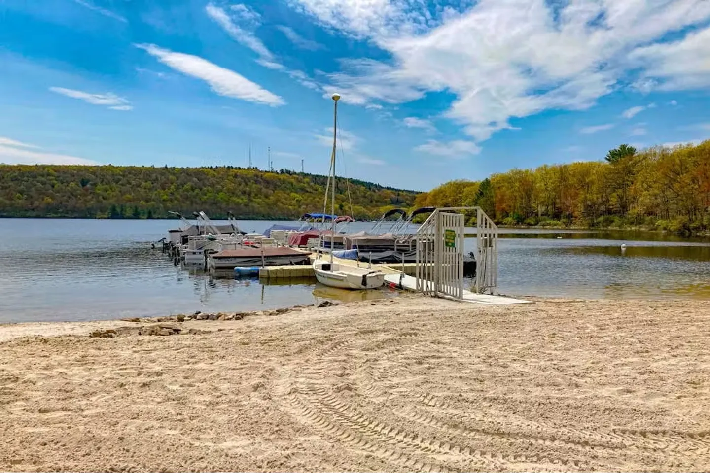 Sandy lake beach leading to a floating dock filled with pontoon boats, with forested hills across the water under a partly cloudy sky.