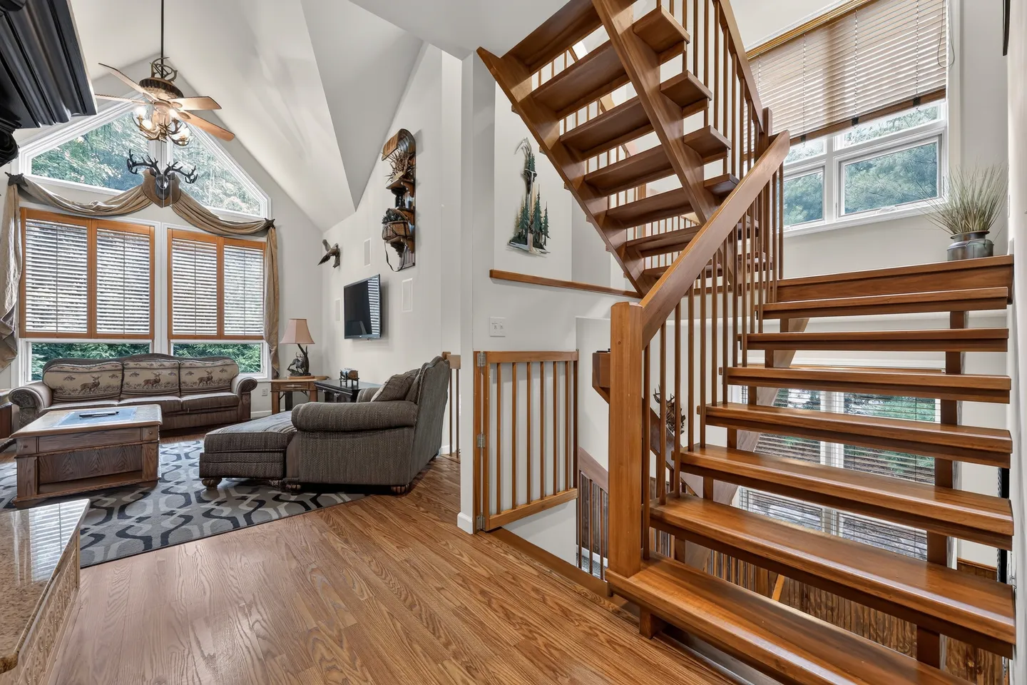 Open wooden staircase next to a living room with vaulted ceilings, large windows, and sofa seating.