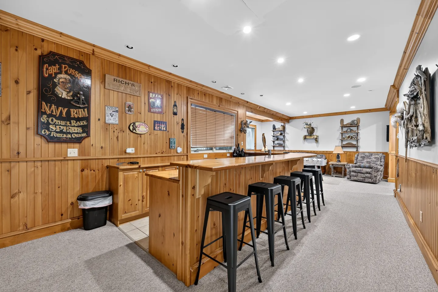 A finished basement featuring a wood-paneled wet bar with black stools, knotty pine walls, and carpeted flooring leading to a sitting area.