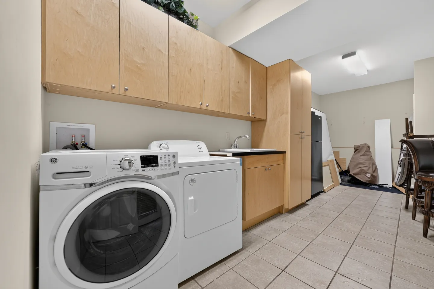 A utility and laundry area featuring a white washer and dryer next to light-wood cabinets, a sink, and tiled flooring.