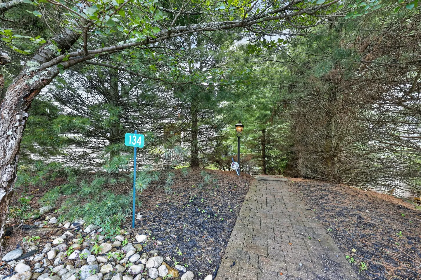 A stone walkway leading through dense evergreen trees past an address post reading 134 and a lamppost.