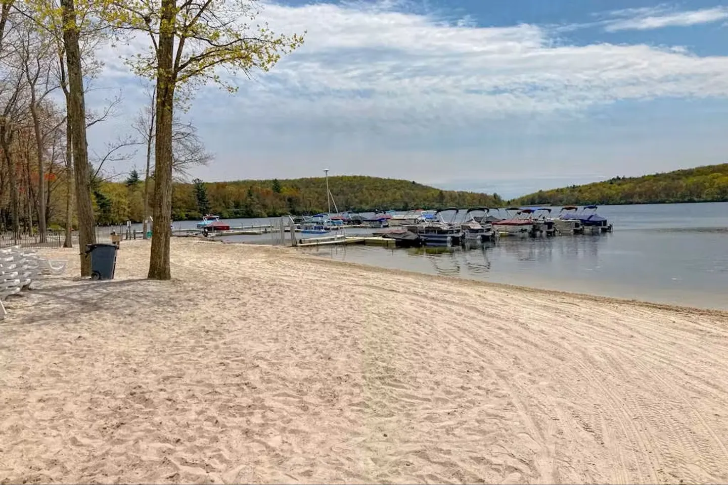 A sandy beach shoreline bordering a calm lake with boat docks and forested hills in the background.