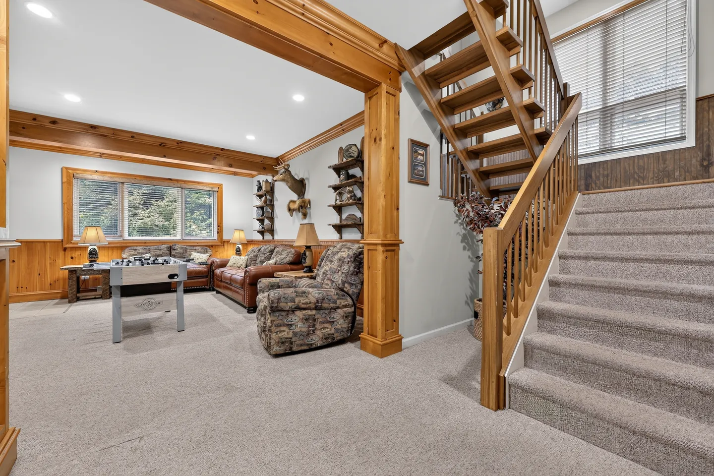 A carpeted lower-level living area viewed from beside a wooden staircase, featuring a foosball table, armchairs, leather sofa, and wood trim.