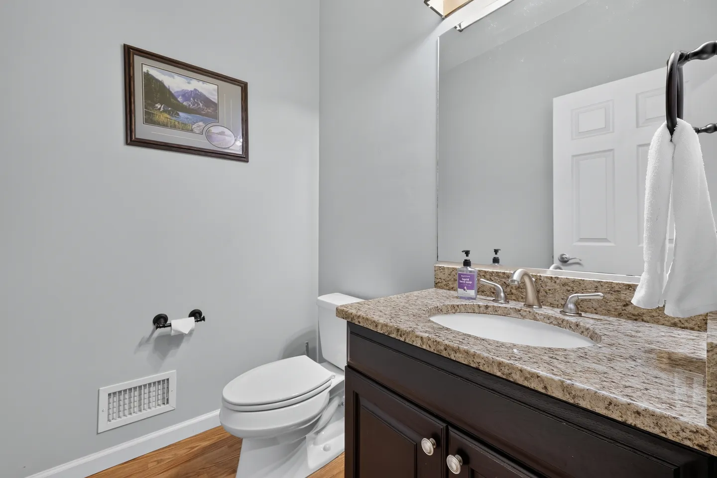 Bathroom with a toilet, wood vanity with granite-style countertop, large frameless mirror, and light gray walls.