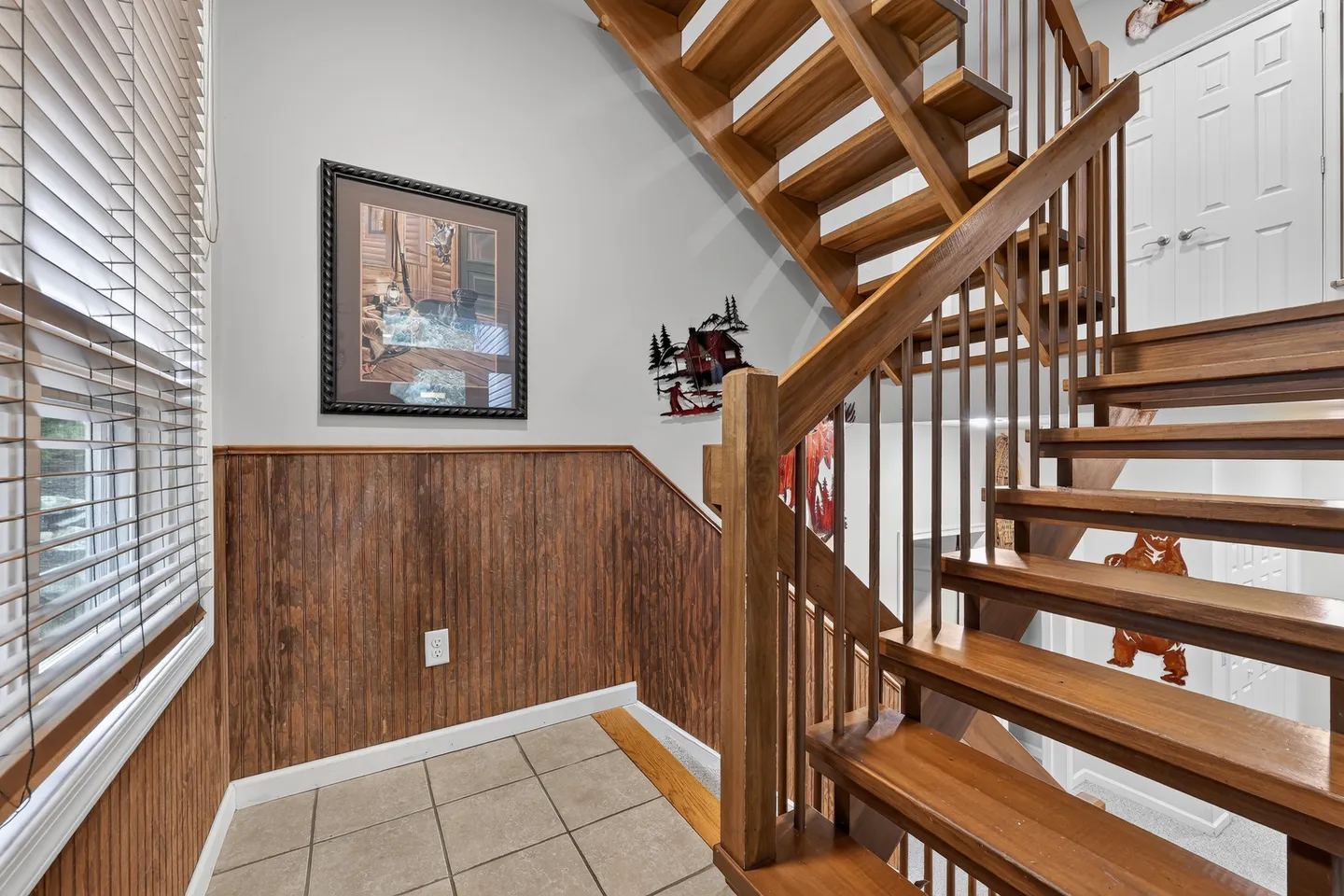 Open-riser wooden staircase with vertical railings beside a tiled foyer with wood wainscoting.