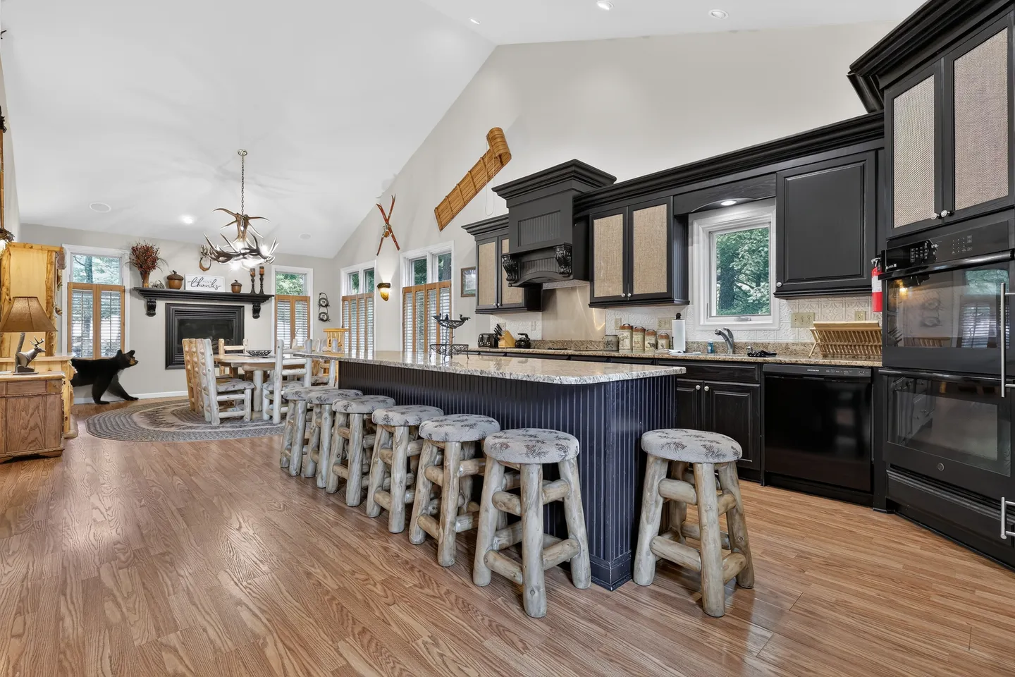 Open-concept kitchen and dining area featuring dark cabinets, a large island with rustic log barstools, and vaulted ceilings.