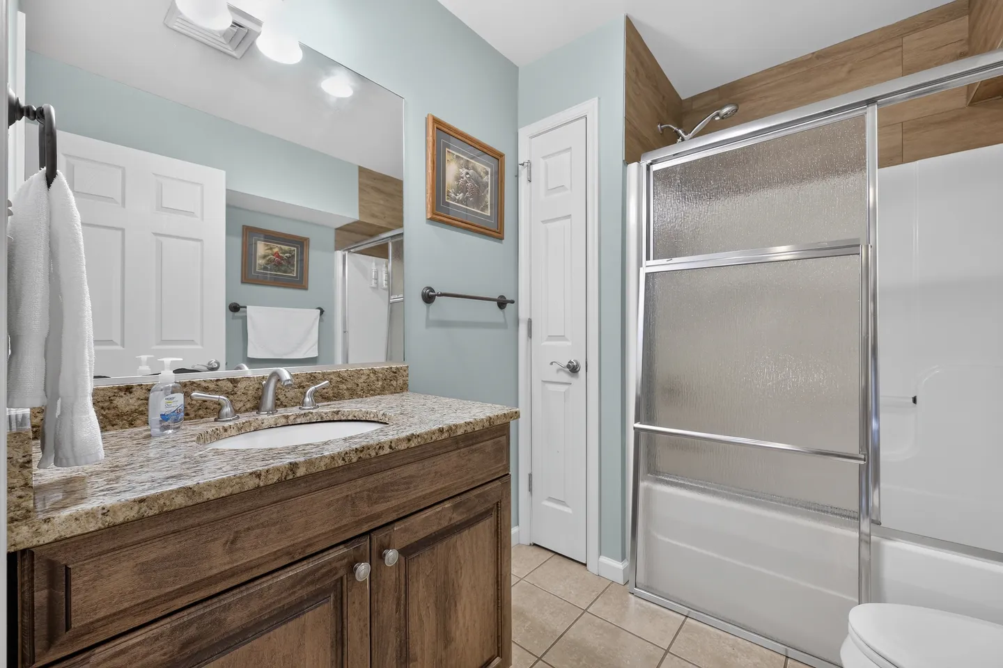Bathroom with a wood vanity, granite-look countertop, large mirror, and a bathtub with textured glass sliding doors.