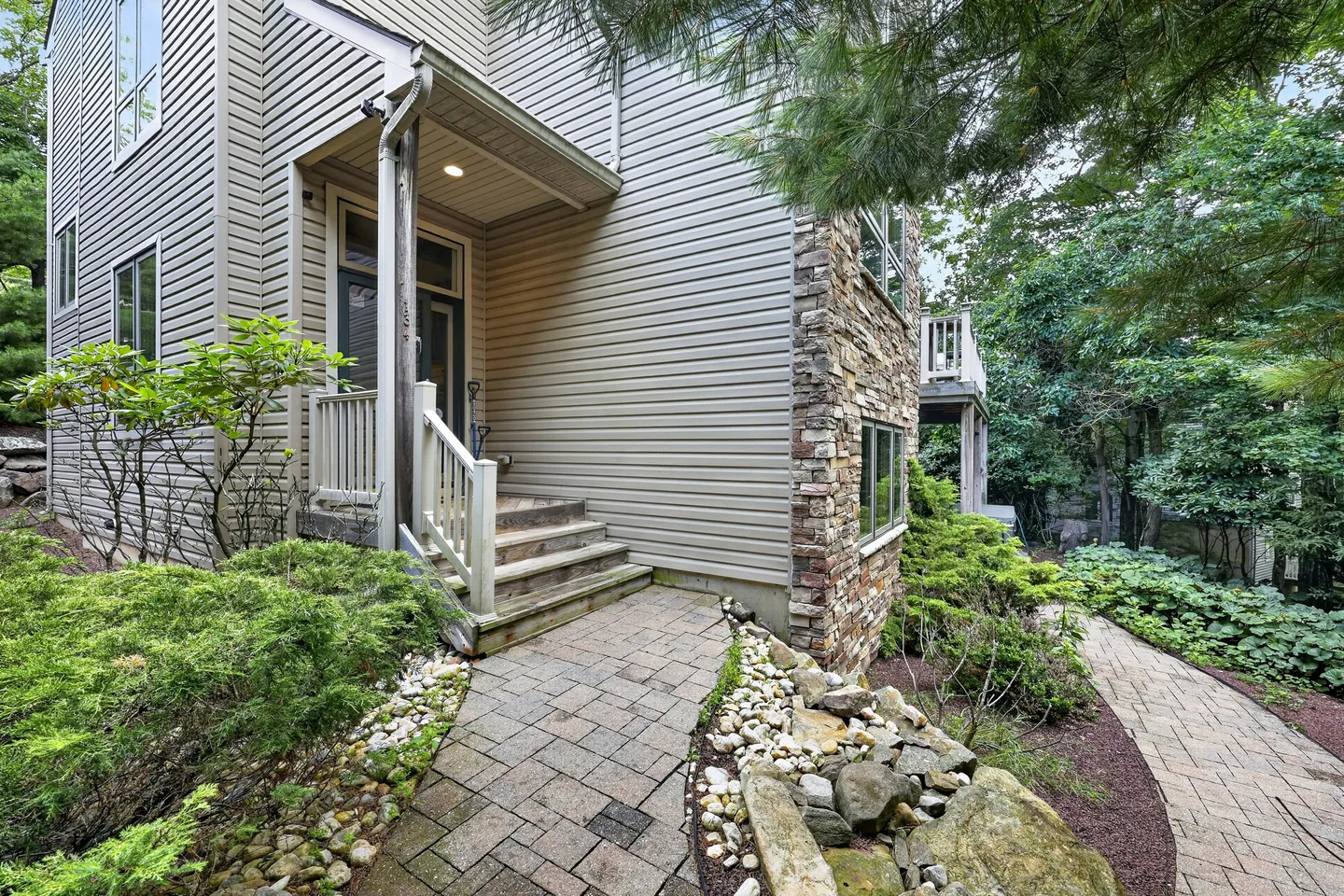 Exterior stone-paved walkway leading to the entrance of a siding and stone-accented home surrounded by trees and landscaping.