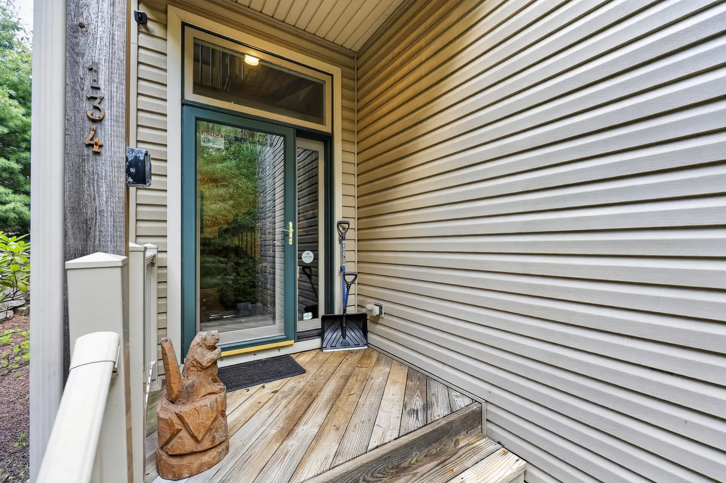 Exterior home entrance with a wooden porch, dark green glass door, light-colored horizontal siding, and house numbers 134 on a post.