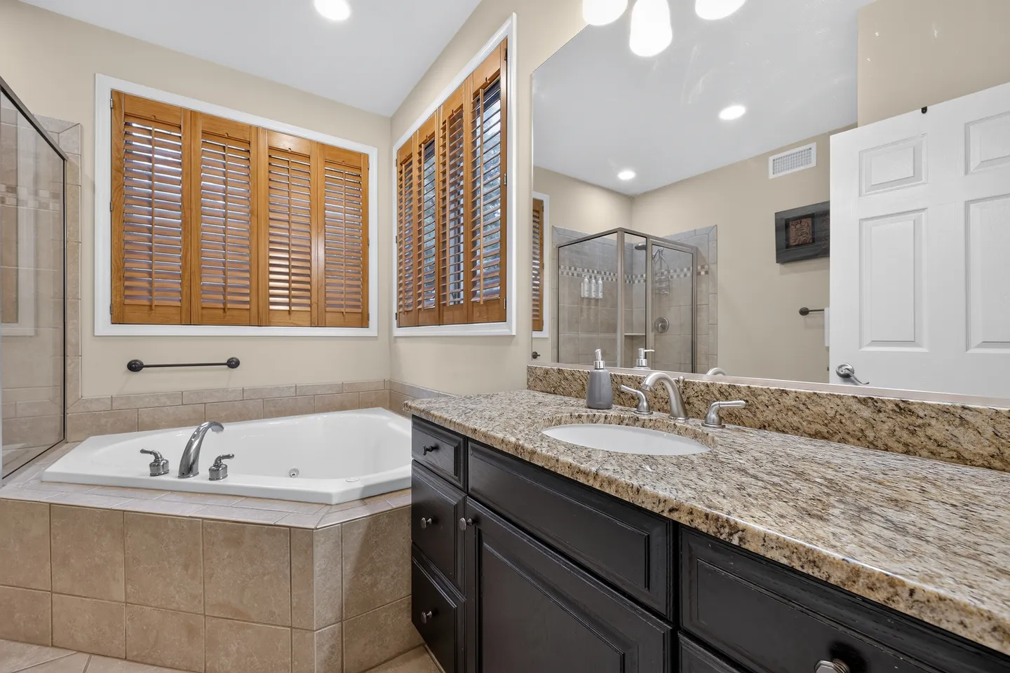 Bathroom featuring a corner soaking tub with tiled surround, a dark vanity with a speckled countertop, and windows with wooden plantation shutters.