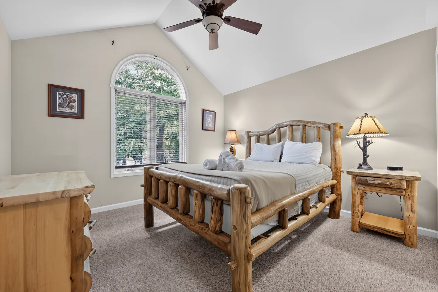 Bedroom with vaulted ceiling, arched window, log-style bed frame, matching nightstand, and ceiling fan.