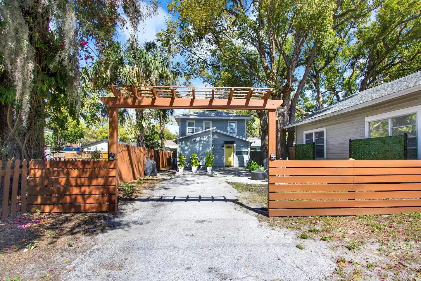 A driveway entrance framed by a wooden pergola and horizontal slat fence leading to a blue house with a yellow front door.