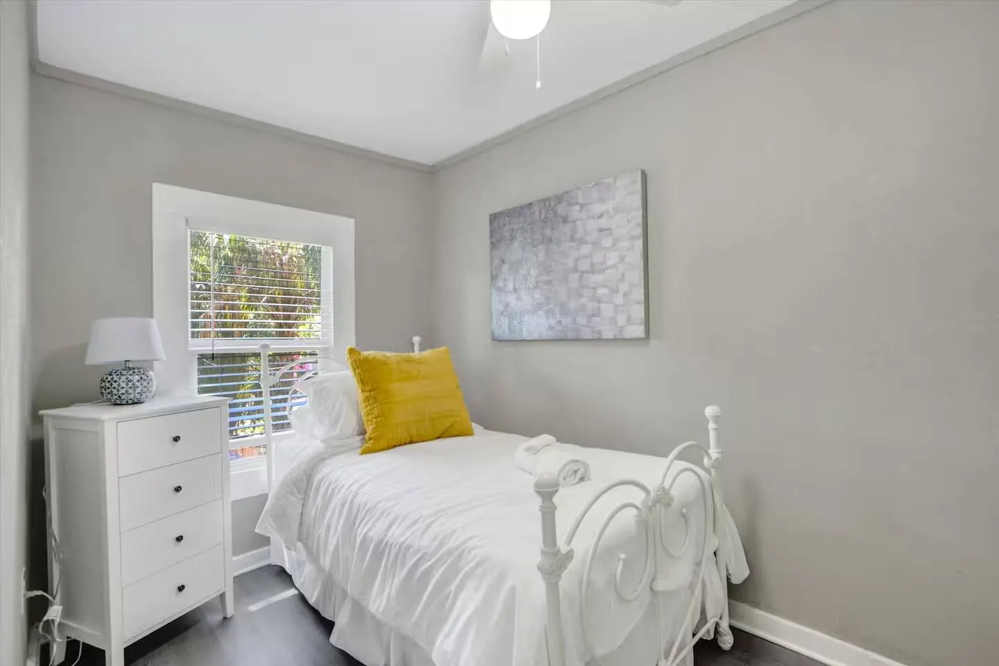 Bedroom with a white metal twin bed, yellow accent pillow, white nightstand dresser, and window with blinds.