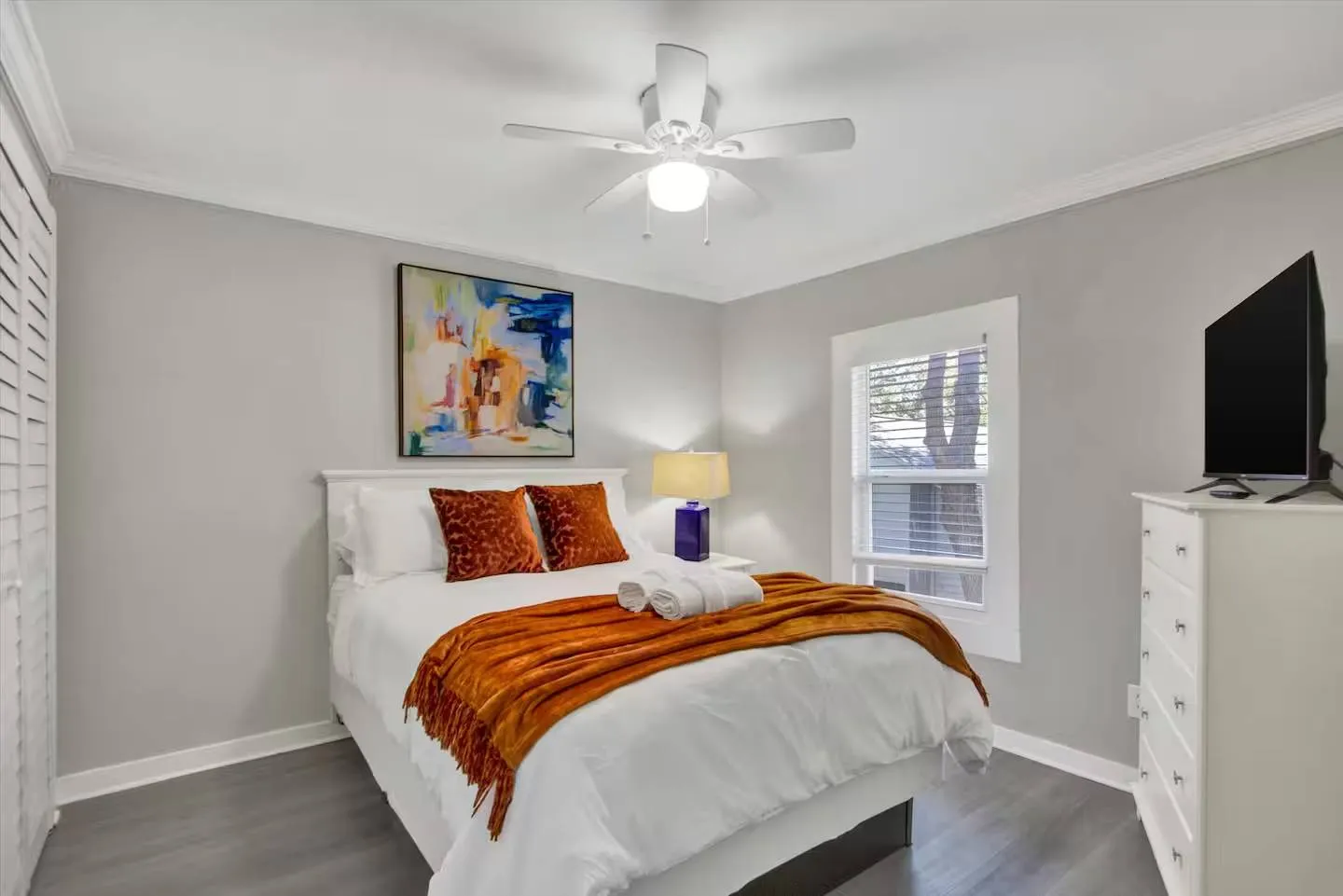 Bedroom featuring a made bed with white linens and orange accents, a ceiling fan, a window, and a mounted TV on a white dresser.