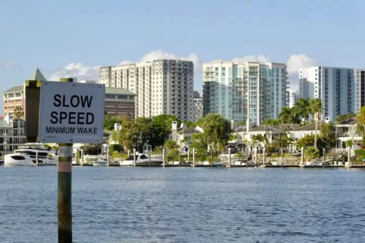 View across a waterway toward residential buildings, boats at docks, and a slow speed minimum wake sign.