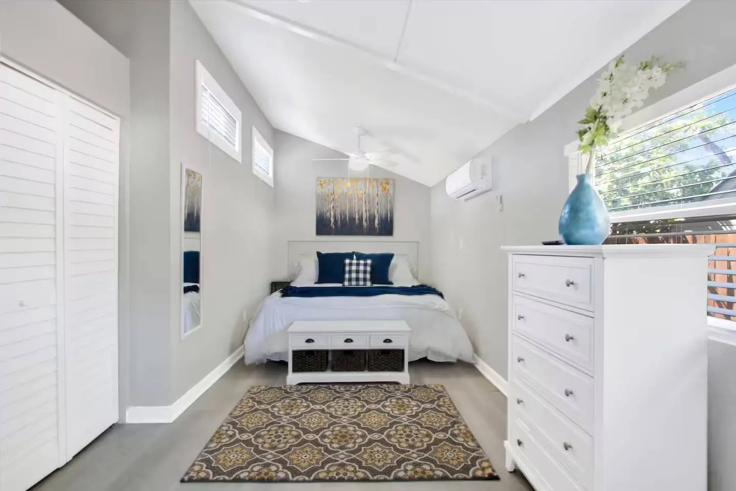 Bedroom with vaulted ceiling, made bed, patterned area rug, white dresser, and wall-mounted AC unit.