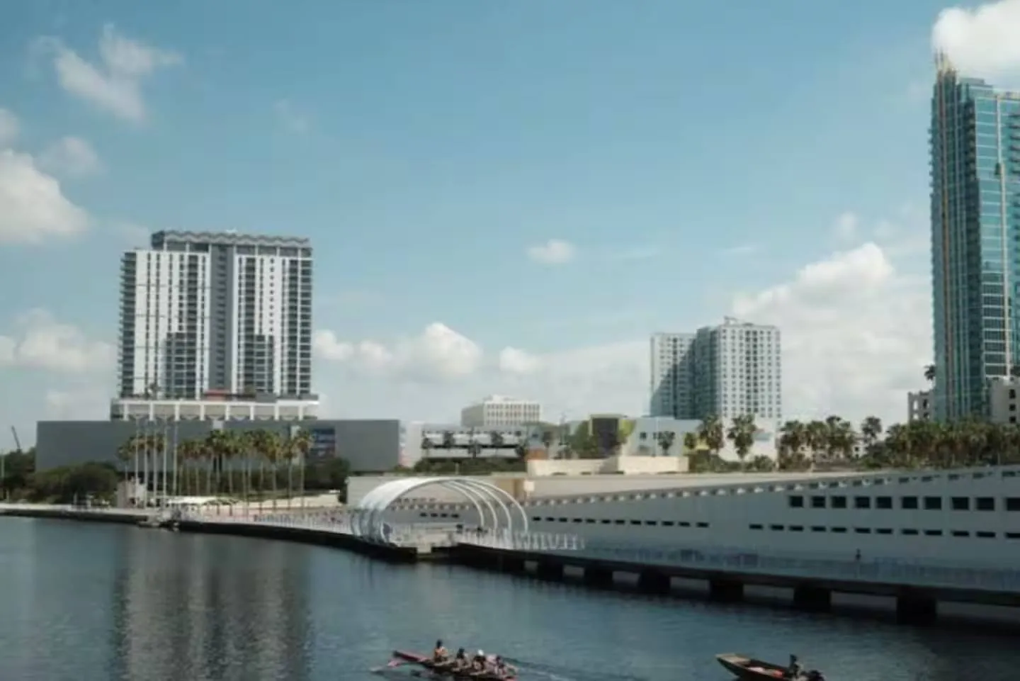 Riverfront view showing an arched bridge structure, riverwalk, rowers on the water, and city high-rises under a blue sky.