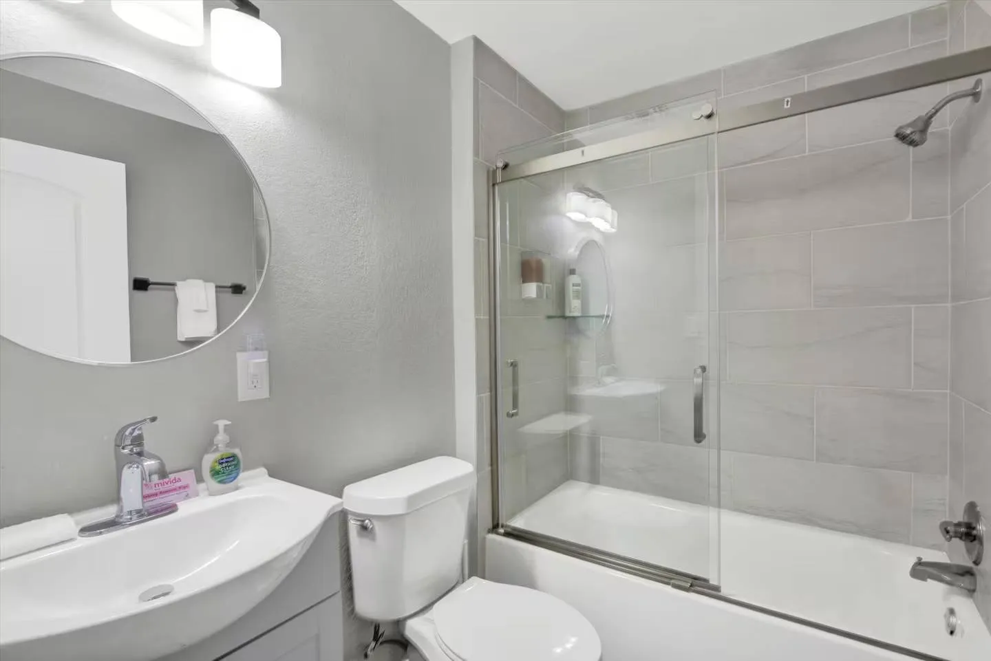Bathroom with a vanity sink, round mirror, toilet, and a glass-enclosed bathtub and shower combination with tiled walls.