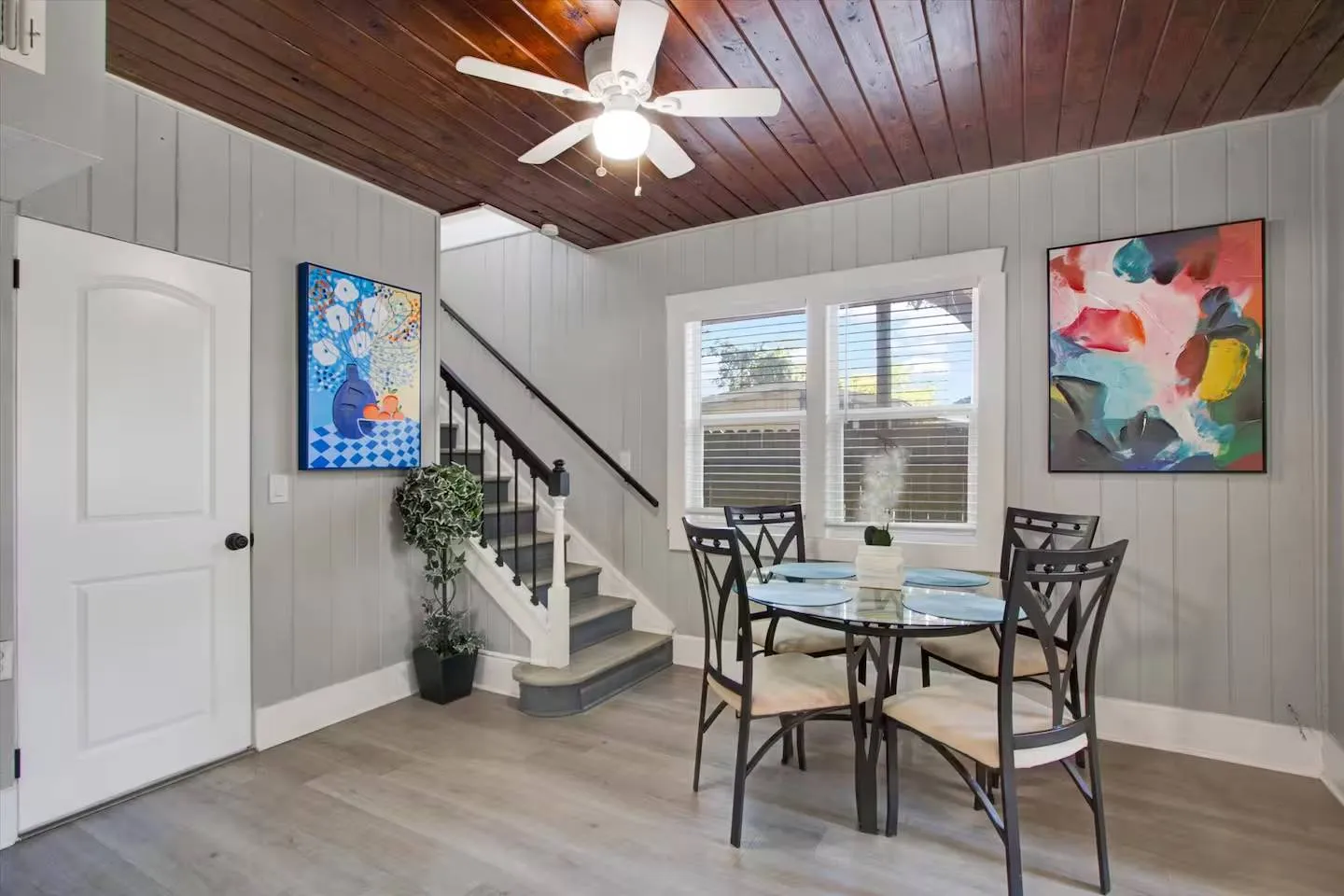 Dining area with a round glass table and four chairs beneath a wood-paneled ceiling, positioned near a staircase and door.