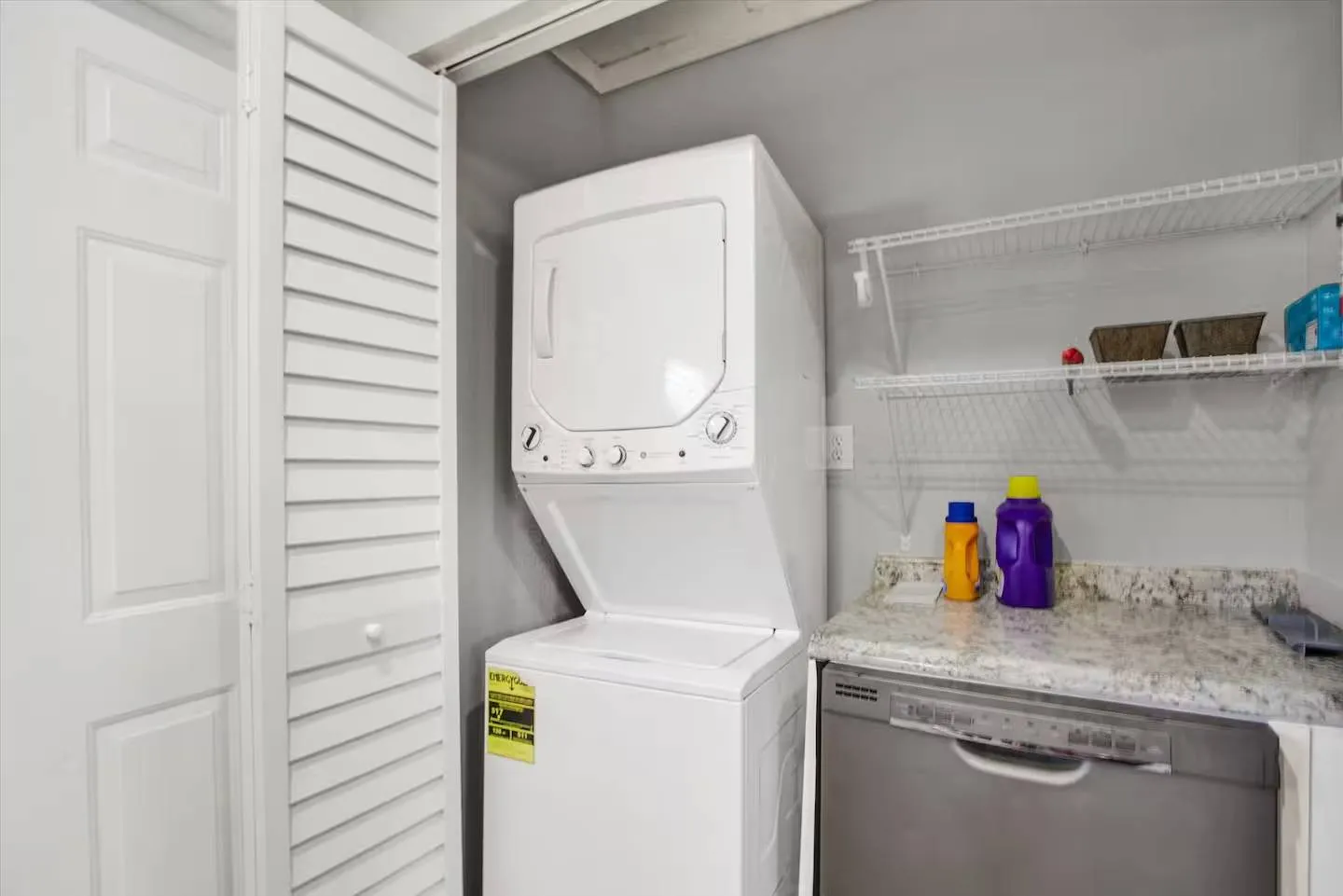 Stacked white washer and dryer unit next to a countertop and wire shelving inside a laundry closet.