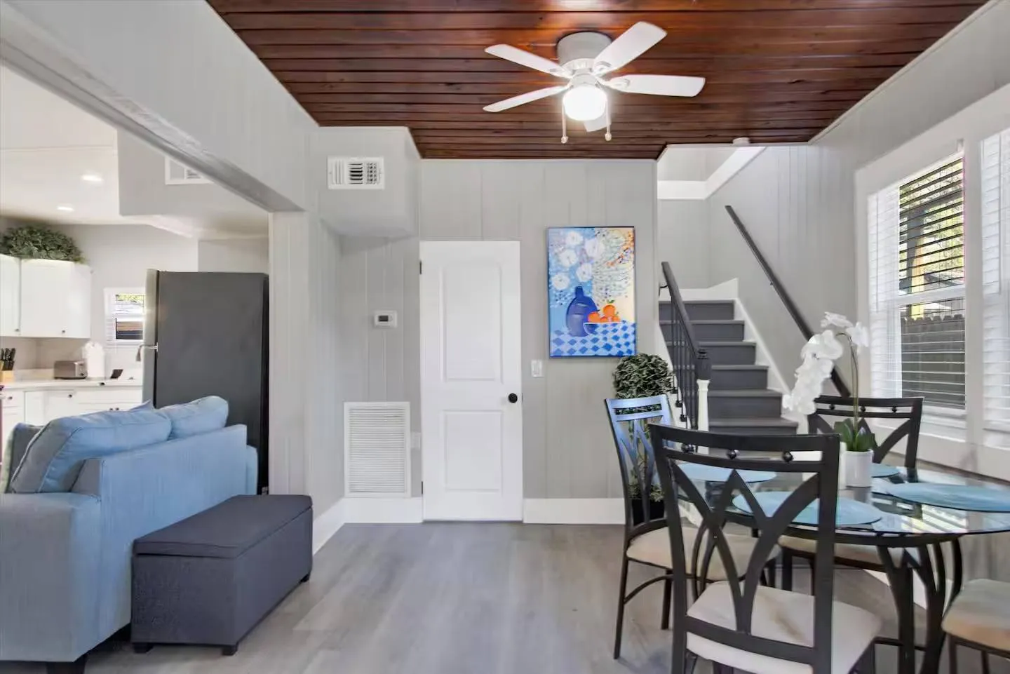 Open living and dining area featuring light gray paneled walls, a dark wood ceiling with a fan, a dining table, stairs, and a doorway to the kitchen.