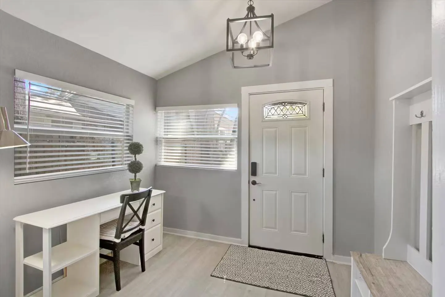 Front entryway with a white front door, light-toned flooring, a modern black chandelier, and a white desk under windows with blinds.