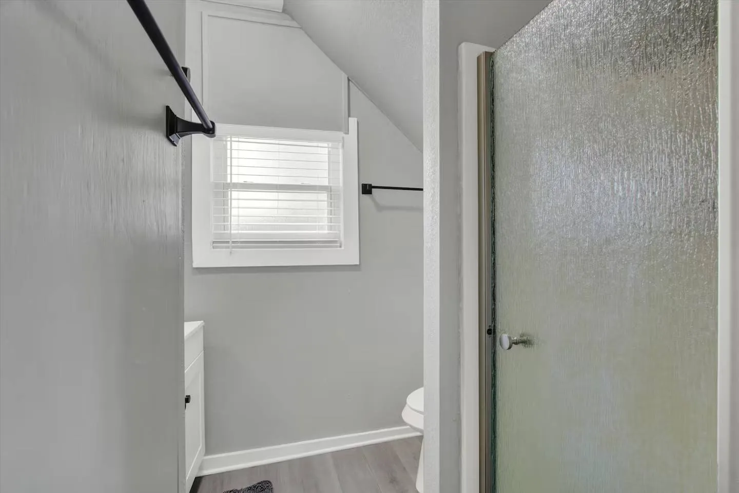 Bathroom with light gray walls, a window with blinds, a toilet under a sloped ceiling, and a textured glass shower door.