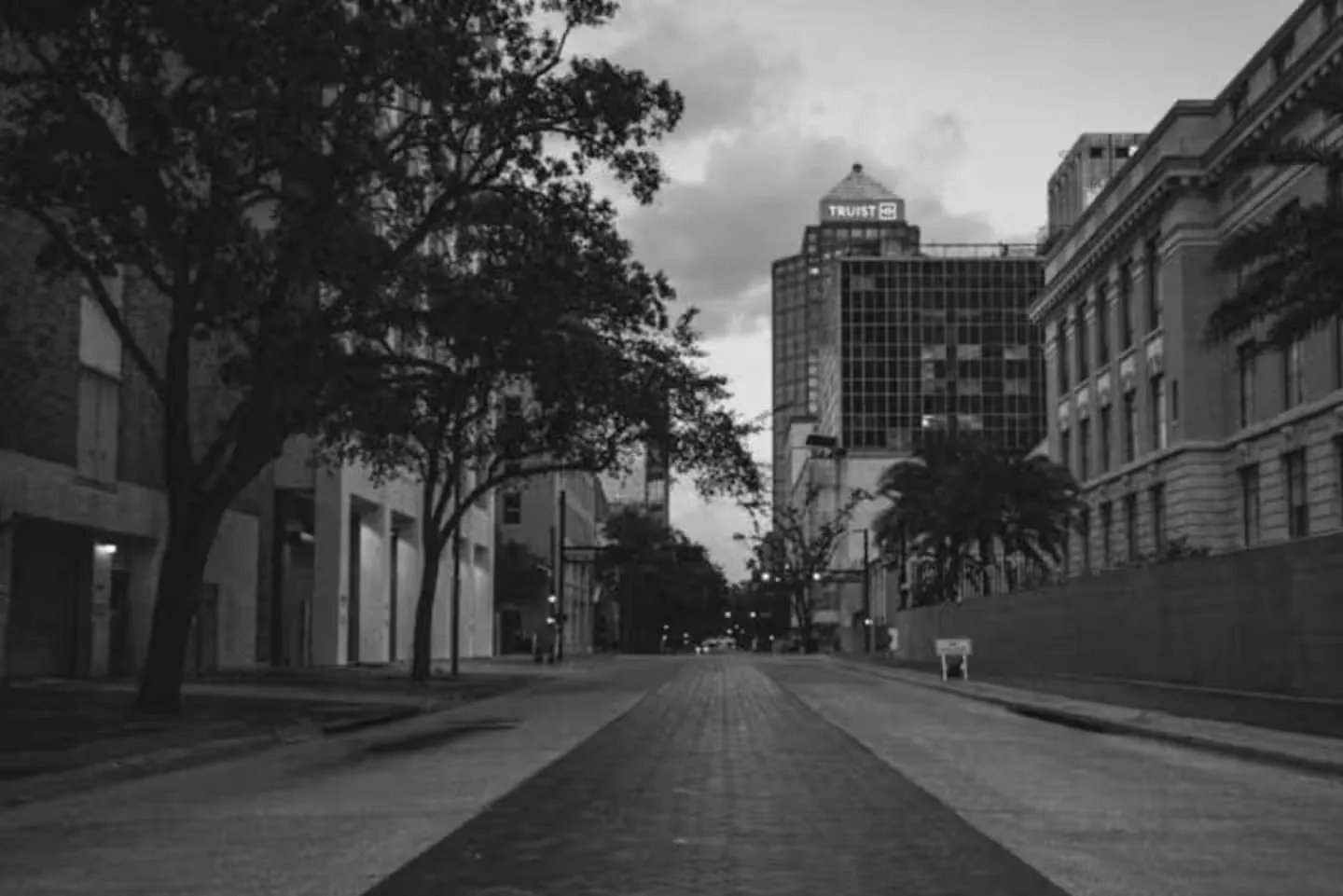 Black and white view looking down a quiet city street lined with trees, sidewalks, and commercial buildings toward a distant high-rise.