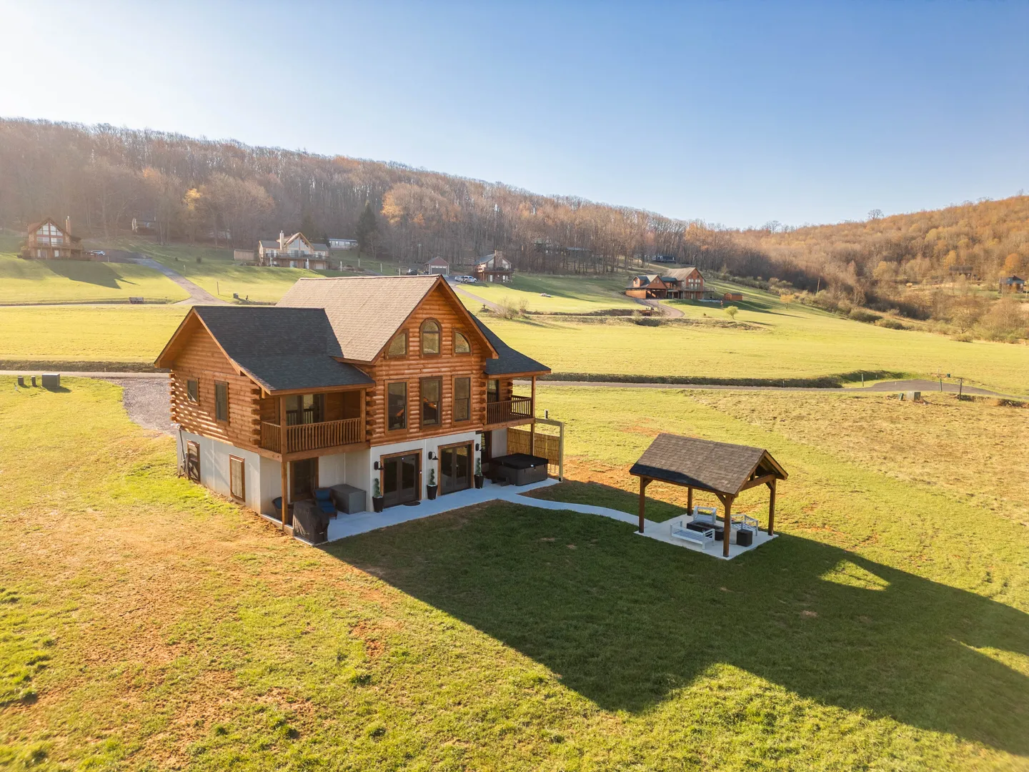 Aerial view of a multi-story log cabin with a covered patio, detached pavilion, and expansive grassy yard against rolling hills.