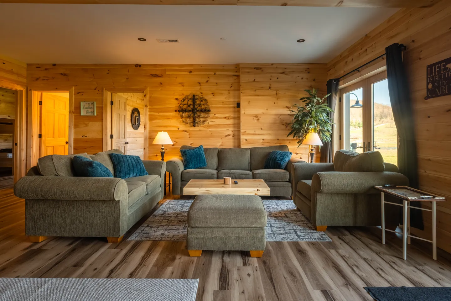 Living room with knotty wood-paneled walls, upholstered seating set with teal accent pillows, and sliding glass doors.