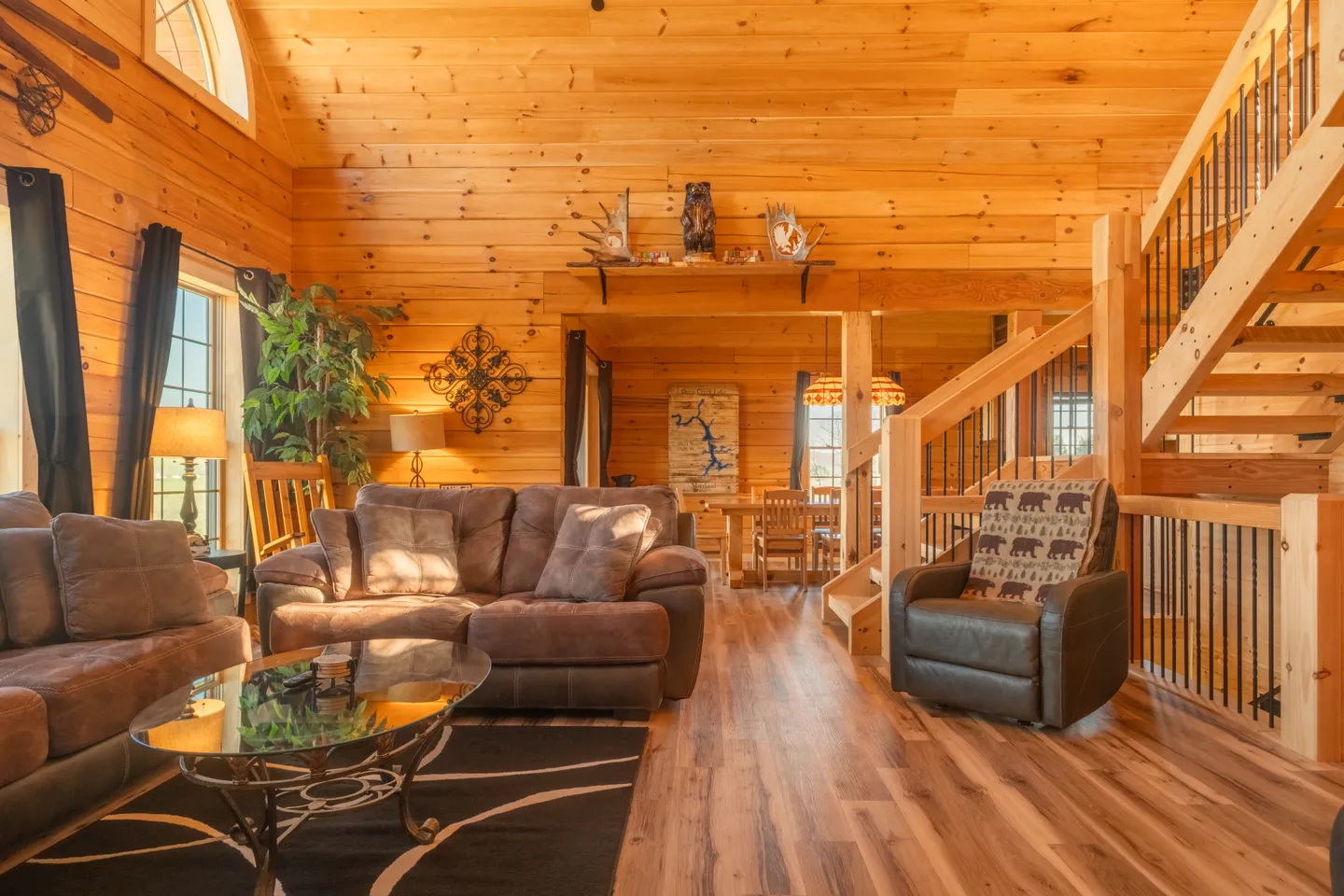 Living room with wood-paneled walls and ceiling, plush brown couches, and a wooden staircase leading upstairs.