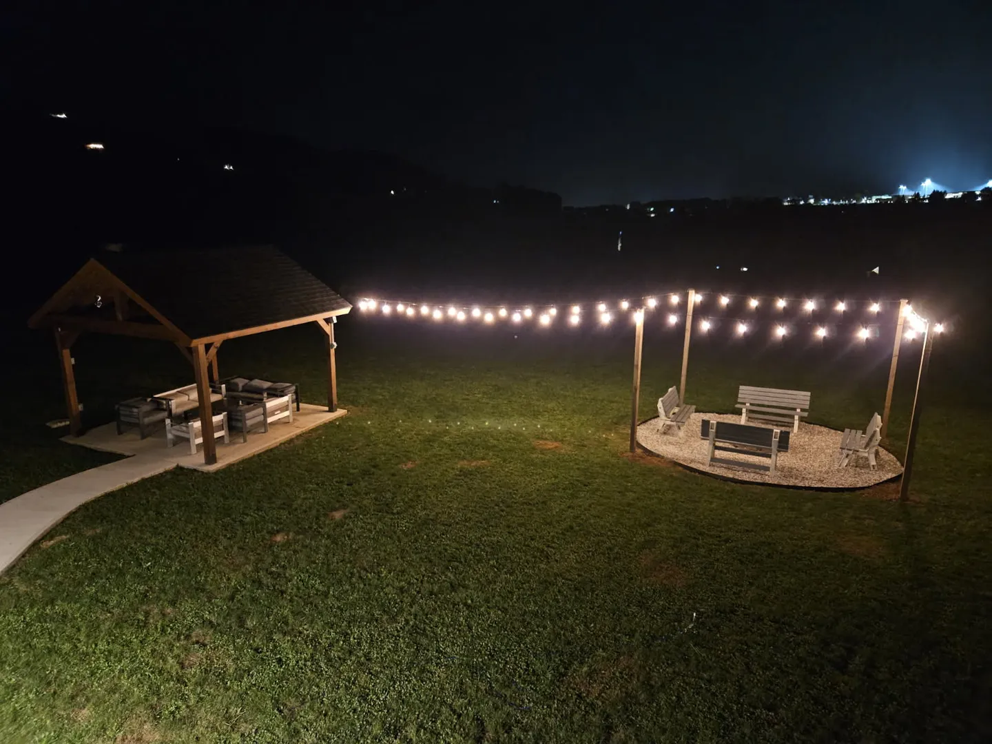 Nighttime view of a grassy yard with a covered pavilion with outdoor seating and a fire pit area surrounded by benches, connected by string lights.