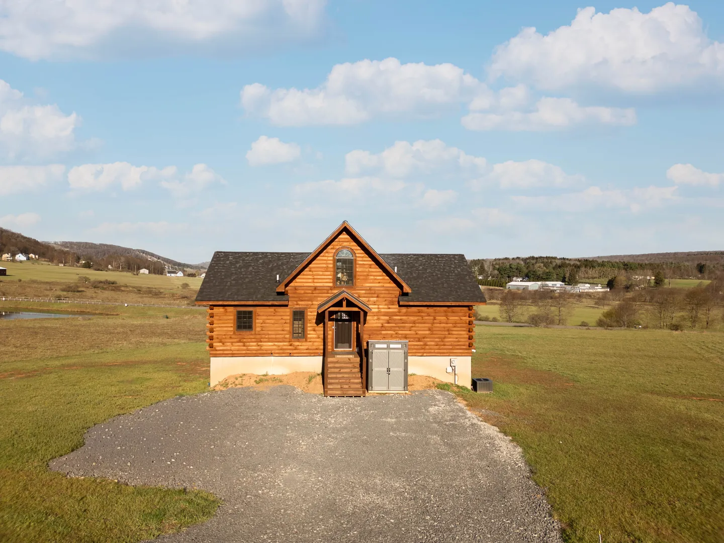 Front exterior view of a log cabin with a pitched dark shingle roof and front steps set in an open grassy field.