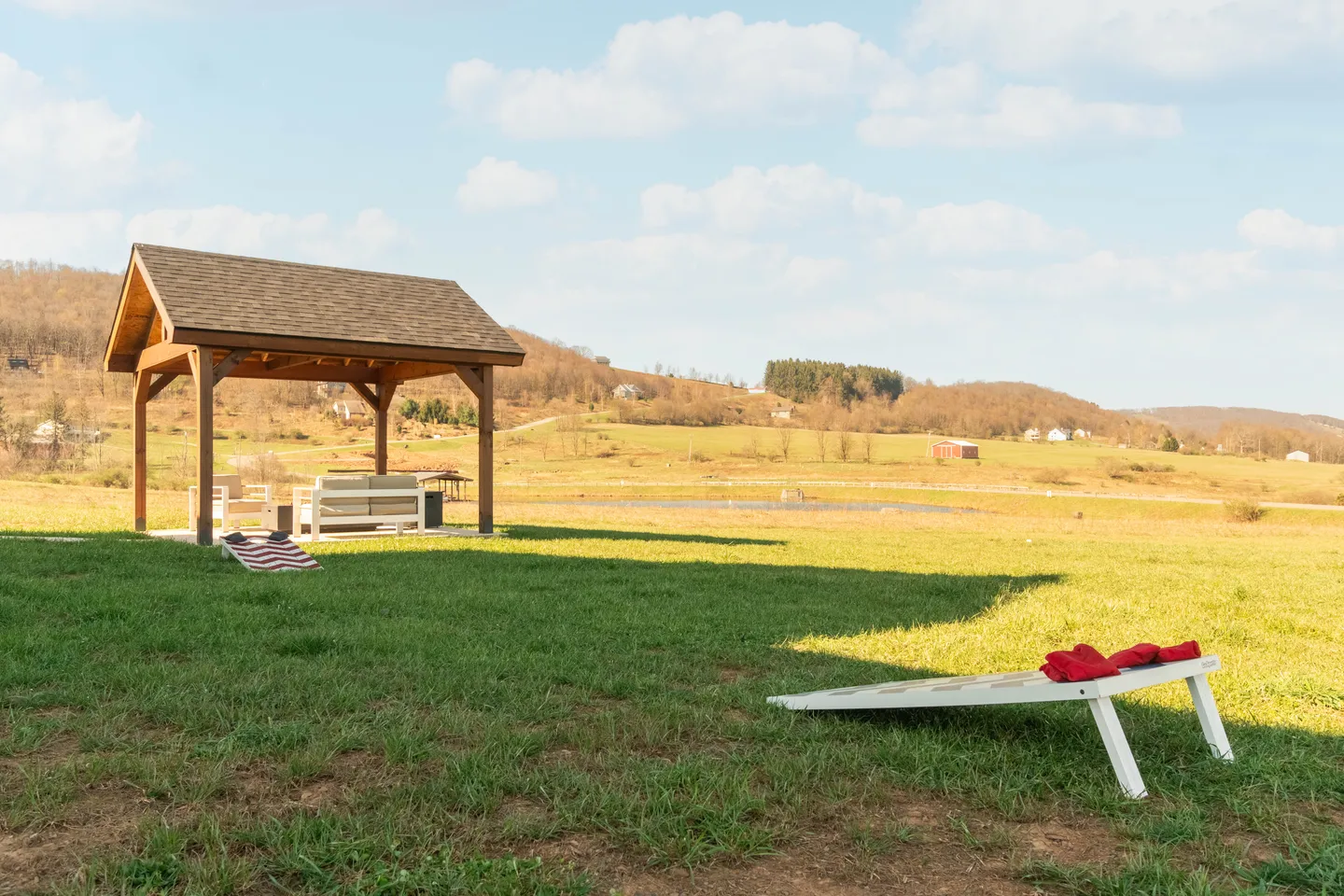 Open grassy yard with a cornhole board in the foreground and a covered wooden pavilion with seating in the background overlooking rolling hills.