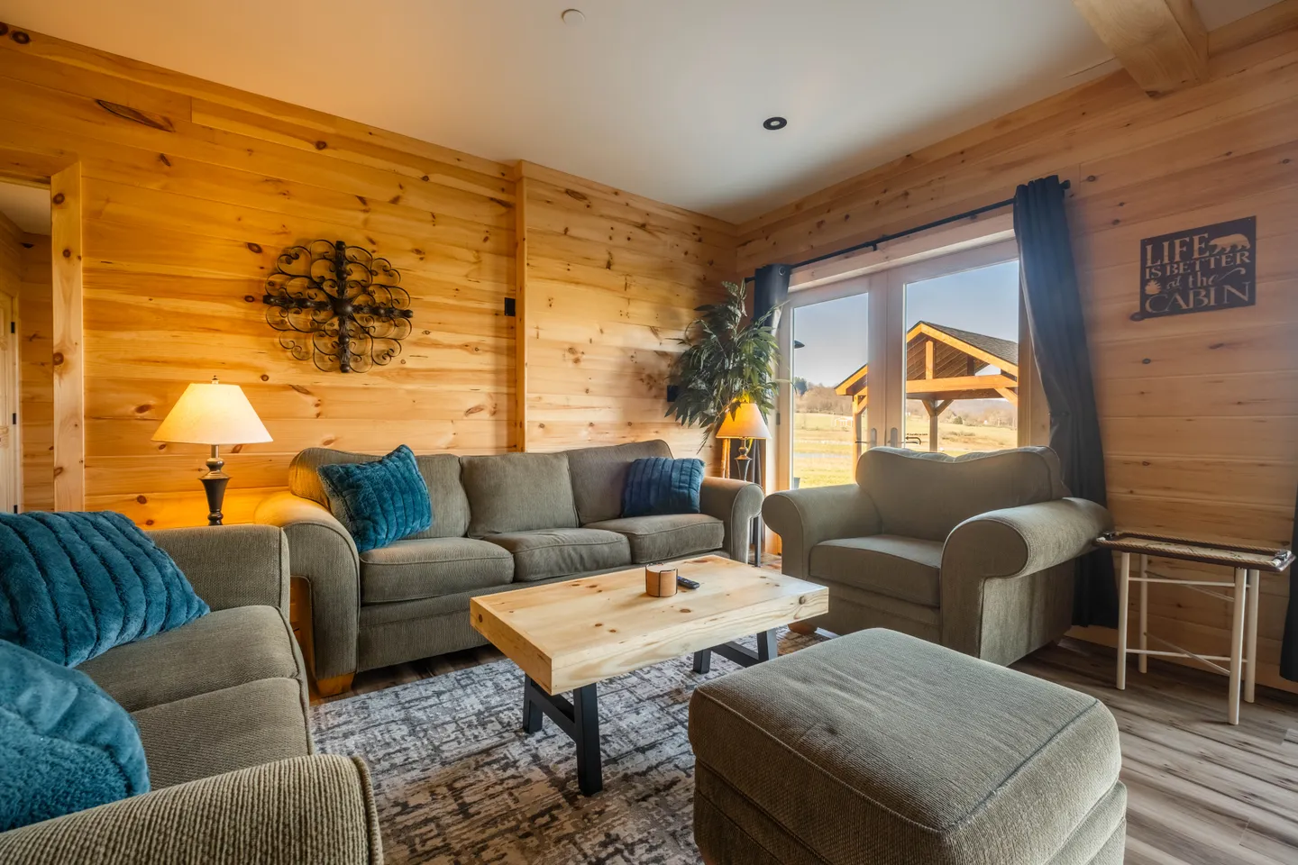 Living room with knotty wood-paneled walls, upholstered seating, a wooden coffee table, and glass doors looking outside.