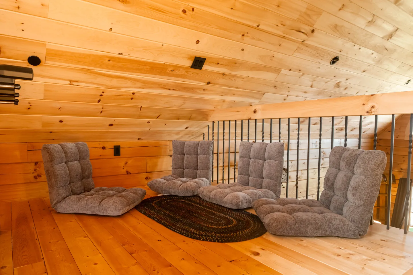 Low-ceiling loft with wood-paneled walls and ceiling, featuring four adjustable floor chairs around a small oval rug next to a railing.