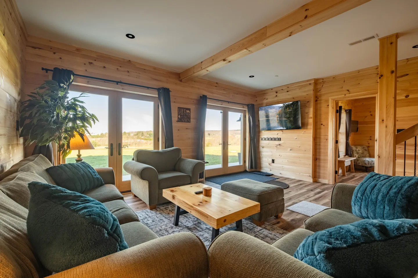Living room with wood-paneled walls, exposed ceiling beams, a sectional sofa, armchair, and double glass doors overlooking a grassy yard.