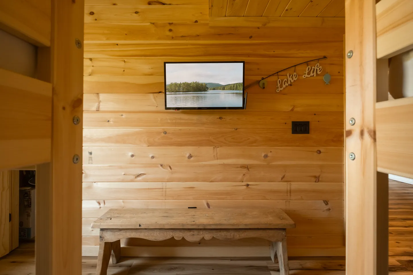 A wooden paneled alcove or room featuring a wall-mounted TV, a rustic bench below, and a 'Lake Life' sign.