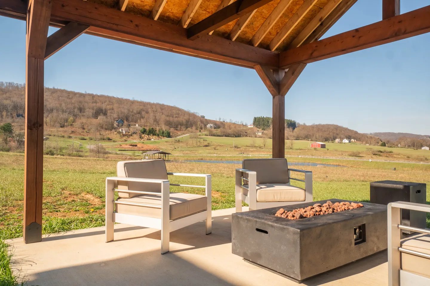 A covered outdoor patio with a fire pit table, armchairs, and scenic view of rolling hills.