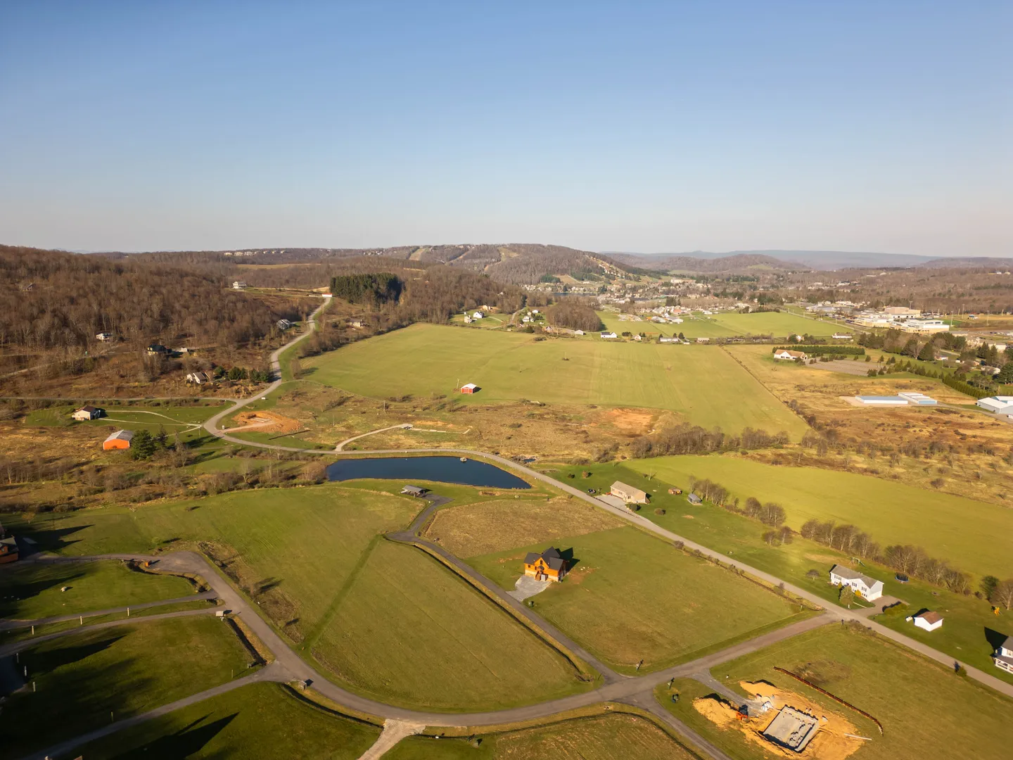 Aerial view of a rural landscape with scattered homes, open green fields, a small pond, and rolling hills under a clear blue sky.