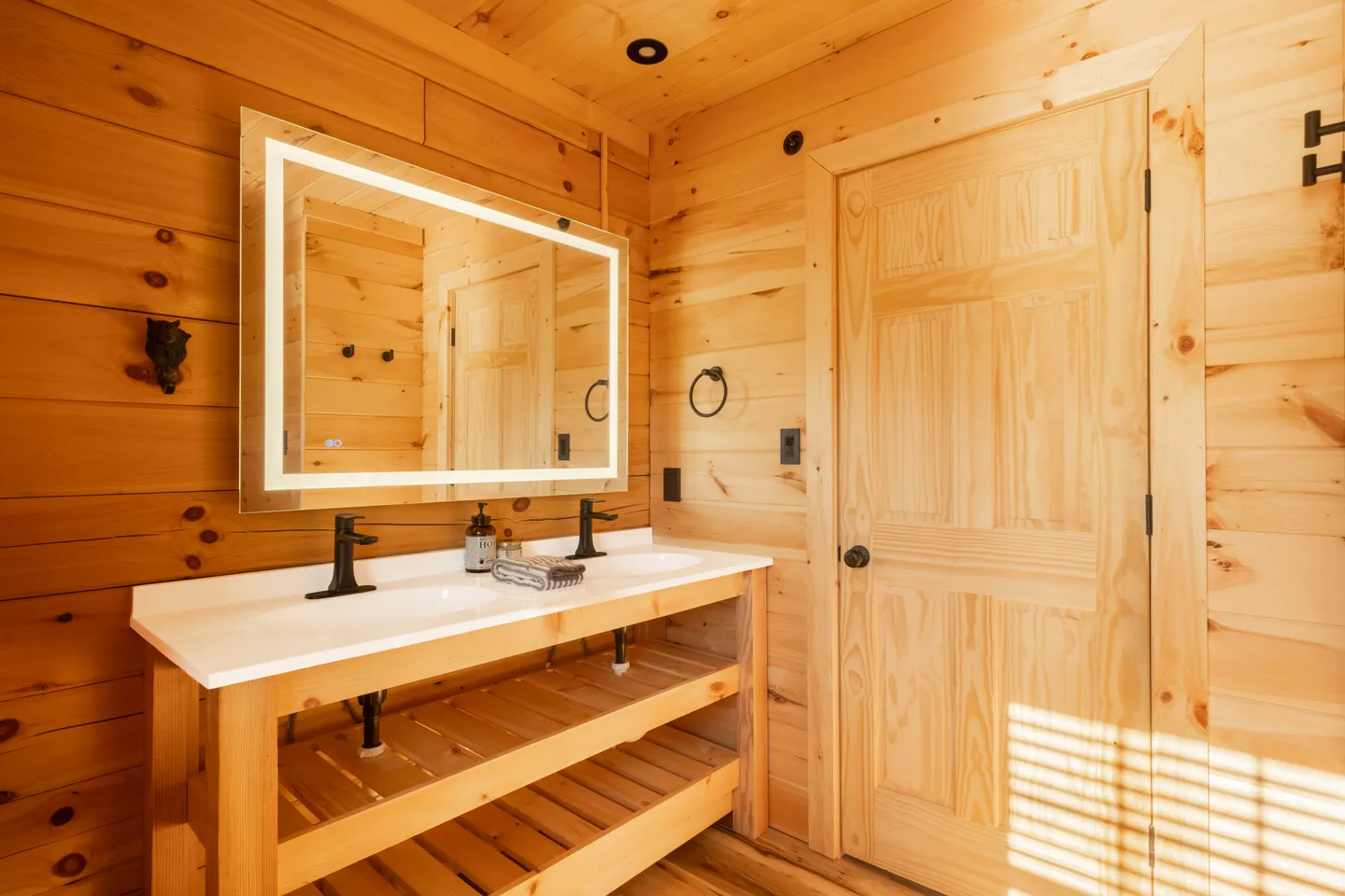 Bathroom with wood-paneled walls, a double vanity with open slatted shelves, and an illuminated rectangular mirror.