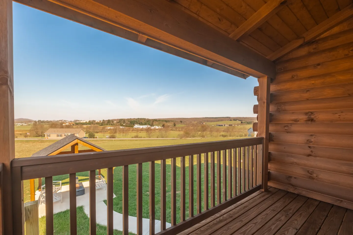 View from a covered wooden porch looking out over green lawn toward distant fields and buildings under a clear blue sky.