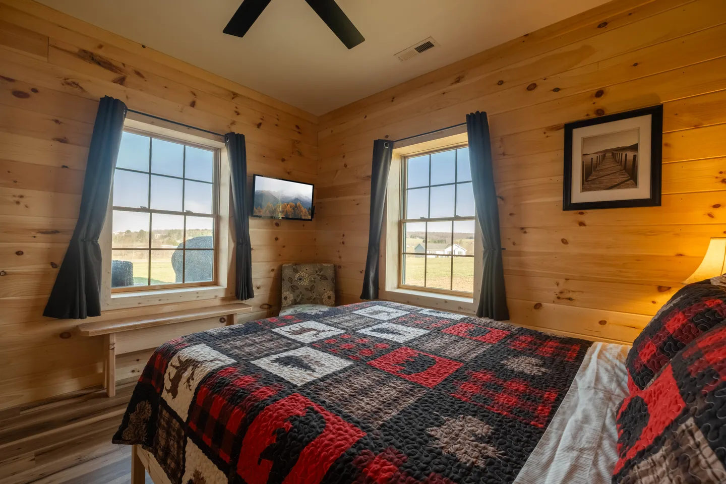 Bedroom with knotty pine wood-paneled walls, a bed with a rustic patchwork quilt, and two windows overlooking fields.