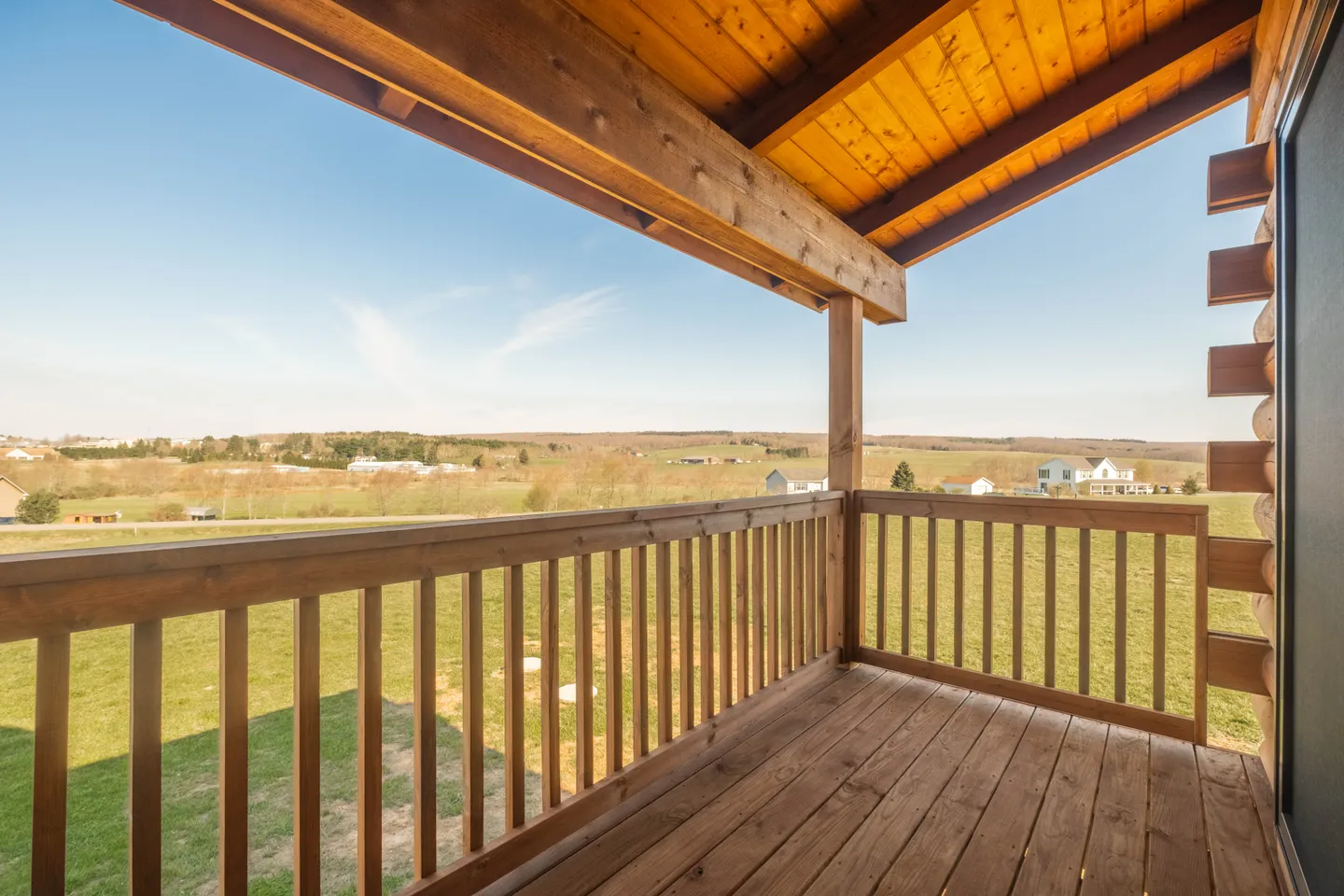 A covered wooden balcony overlooking an open grassy field with rolling hills under a blue sky.