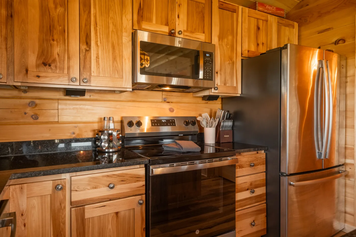 Kitchen area showing stainless steel appliances including a refrigerator, range, and over-the-range microwave set among natural wood cabinets and log walls.