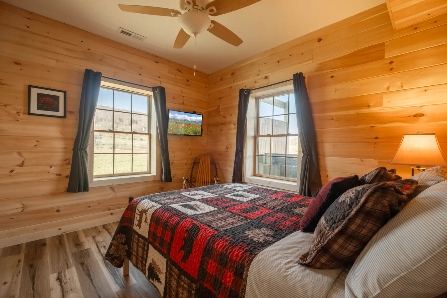 Bedroom with light wood paneled walls, a rustic quilt bedspread, ceiling fan, and two windows showing outdoor views.