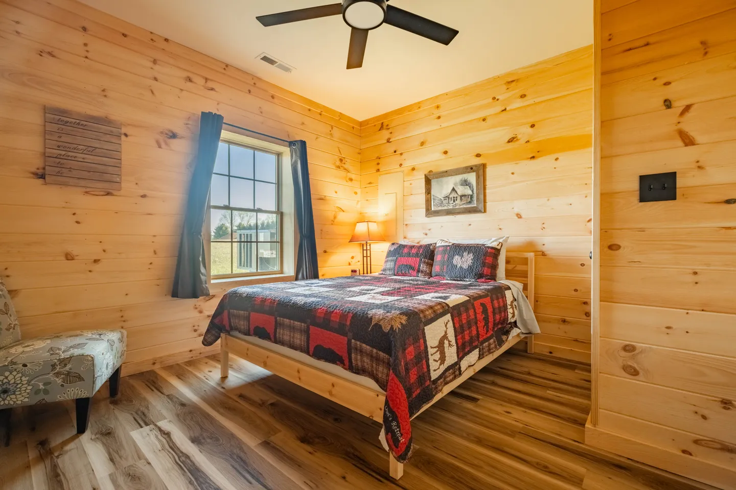 Bedroom with knotty pine wood paneled walls, wood-look flooring, a bed dressed with a rustic patterned quilt, and a ceiling fan.