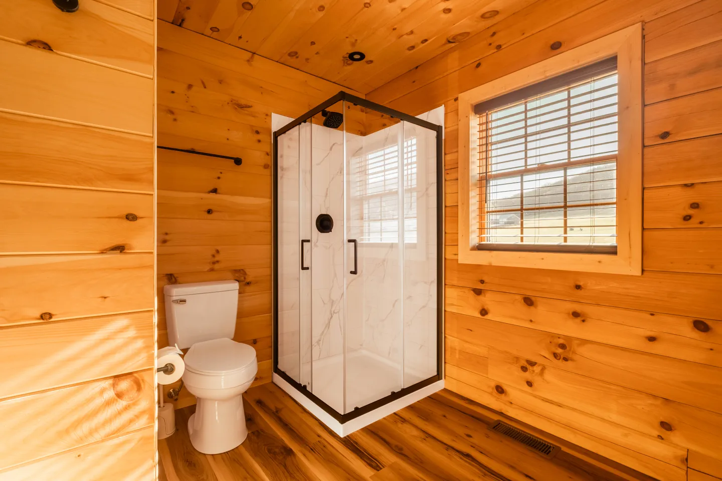 Bathroom with knotty pine wood walls, wood-look flooring, a white toilet, and a corner glass shower stall next to a window with blinds.